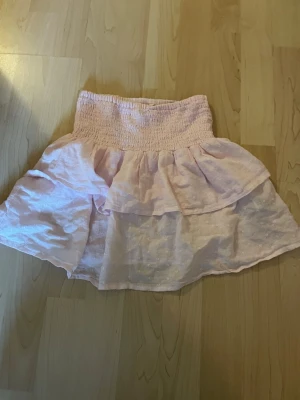 Ljusrosa volangkjol med resår - Söt ljusrosa kortkjol med volang och bred resår i midjan. Kjolen har en luftig och lätt känsla, perfekt för varmare dagar. Materialet ser ut att vara bomull eller en bomullsblandning. Enkel att matcha med olika toppar.