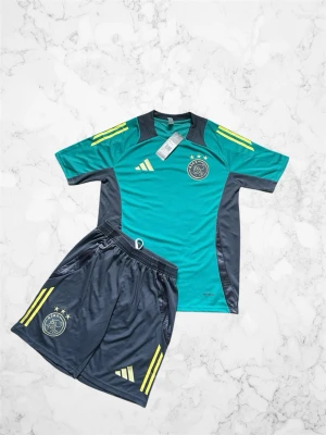 Ajax träningsset i turkos och mörkgrå - Ajax träningsset med t-shirt och shorts i turkos och mörkgrått. Setet har gula detaljer och klubbmärke på både tröja och shorts. Tillverkat i ett lätt och ventilerande material, perfekt för fotbollsträning eller sportaktiviteter.