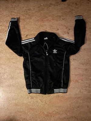 Adidas track jacket - Svart track jacket från Adidas med vita ränder längs ärmarna och vita detaljer. Dragkedja framtill, ribbade muddar och krage. Adidas logga på bröstet och stort vitt Trefoil-tryck på ryggen. Klassisk sportig stil i glansigt material. Perfekt för dig som gillar streetwear/vintage/Y2K. Bara skriva om ni vill ha mått.😊📏