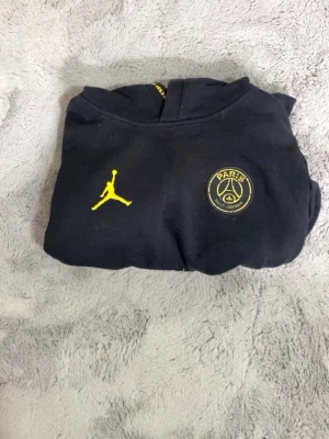 Svart PSG x Jordan hoodie (12-13år) - Snygg svart hoodie med gult PSG x Jordan-tryck både fram och stort på ryggen. Hoodien har klassisk passform och är perfekt för dig som gillar streetwear och sportmode. Materialet känns mjukt och bekvämt. Ikoniska loggor för Paris Saint-Germain och Jumpman.