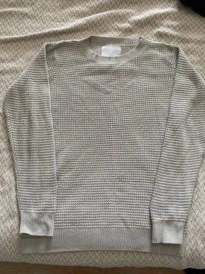 Beige stickad tröja från 157 - Snygg beige stickad tröja från 157 med rund halsringning och diskret strukturmönster. Tröjan har långa ärmar och ribbade muddar vid ärmslut och nederkant. Perfekt för lager-på-lager och enkel att matcha med jeans eller kjol.
