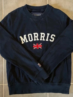 Morris  - Mörkblå sweatshirt från Morris med vit broderad logga och brittisk flagga på bröstet. Tröjan har rund halsringning, långa ärmar och ribbade muddar vid ärmslut och nederkant. Perfekt för en avslappnad och stilren look.