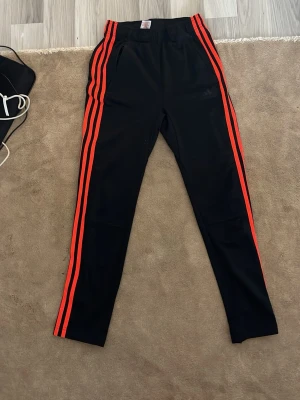 Adidas Svarta mjukisbyxor med röda ränder - Snygga svarta mjukisbyxor med tre röda ränder längs sidorna. Byxorna har smal passform, resår i midjan och sidofickor. Perfekta för en sportig och avslappnad stil. Materialet känns mjukt och stretchigt.