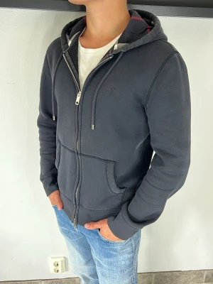 Burberry zip hoodie  - Snygg mörkgrå hoodie med dragkedja och huva. Tröjan har dragsko i huvan, två stora fickor framtill och ribbade muddar vid ärmslut och nederkant. Perfekt för en avslappnad stil och passar till jeans eller joggers.