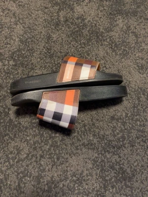 Burberry sandaler med rutigt mönster - Snygga sandaler från Burberry med klassiskt rutigt mönster i orange, brunt, svart och vitt. Remmen är bred och fotbädden är svart med Burberry-logga. Perfekta för en trendig och avslappnad stil.