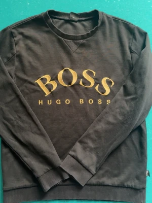 Svart sweatshirt från Hugo Boss - Svart sweatshirt från Hugo Boss med stort broderat BOSS-tryck i gult på bröstet. Klassisk rund halsringning och ribbade muddar vid ärmslut och nederkant. Perfekt för en stilren och avslappnad look.