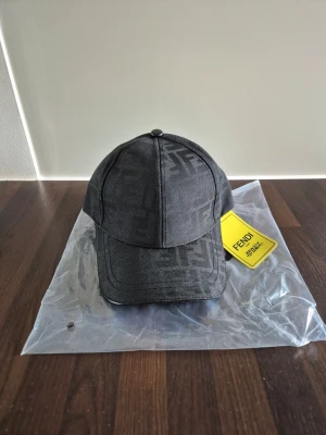 Fendi keps - Snygg svart keps från Fendi med det klassiska FF-monogrammet ton-i-ton över hela kepsen. Kepsen har böjd skärm och justerbar rem bak. Tillverkad i ett slitstarkt tyg som ger en exklusiv känsla. Perfekt accessoar för dig som gillar streetwear och designerplagg.
