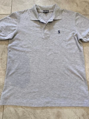 Ljusgrå pikétröja från Polo Ralph Lauren - Klassisk ljusgrå pikétröja från Polo Ralph Lauren med blå broderad logga på bröstet. Tröjan har krage, knappar vid halsen och korta ärmar. Perfekt för en avslappnad men stilren look.
