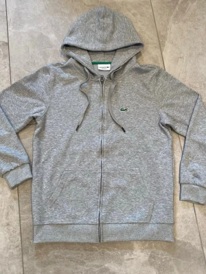Grå zip hoodie från Lacoste - Snygg grå hoodie från Lacoste med dragkedja framtill och klassisk krokodillogo på bröstet. Regular fit-modell med huva och dragsnören samt två fickor framtill. Tillverkad i mjukt material som känns skönt mot huden.