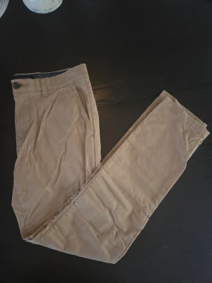 Beige chinos från Tommy Hilfiger 30/32 - Snygga beige chinos från Tommy Hilfiger i slim fit-modell. Byxorna har klassiska fickor fram och bak, bälteshällor och stängs med knapp och dragkedja. Tillverkade i ett mjukt och bekvämt bomullsmaterial som passar perfekt till din vardagsstil.