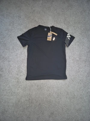 Asics - Svart tränings-tshirt från Asics med rund hals och korta ärmar. Asics-logga på bröstet och vitt tryck på ena ärmen. Tillverkad i ett lätt och snabbtorkande funktionsmaterial som passar perfekt till träning.
