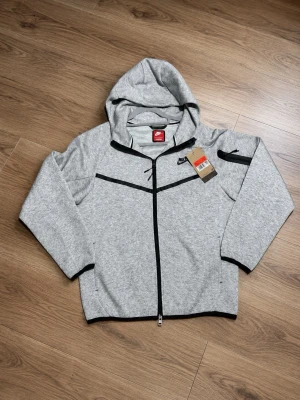 Nike tech fleece M - Snygg grå hoodie från Nike med dragkedja framtill och svarta detaljer. Hoodien har huva, två sidofickor och en extra ficka på ärmen. Tillverkad i mjukt material och har en sportig look med Nike-logga på bröstet.