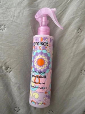 amika brooklyn bombshell blowout spray - Säljer en amika brooklyn bombshell blowout spray i en pastellfärgad flaska med blommigt mönster. Sprayflaskan är rosa och lila med färgglada detaljer och har en praktisk spraypump. Perfekt för att ge volym och glans till håret.