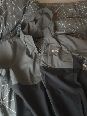 Grå och svart vindjacka Under Armour - Snygg vindjacka från Under Armour i grått och svart med dragkedja och huva. Jackan har en bröstficka med dragkedja och logga på bröstet. Perfekt för blåsiga dagar och sportiga outfits. Lite sönder vid ryggen men lätt att sy 🧵 