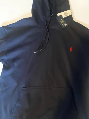 Mörkblå hoodie från Polo Ralph Lauren - Snygg mörkblå hoodie från Polo Ralph Lauren med klassisk röd broderad logga på bröstet. Hoodien har huva med dragsko och en stor magficka. Perfekt för en chill och stilren look.