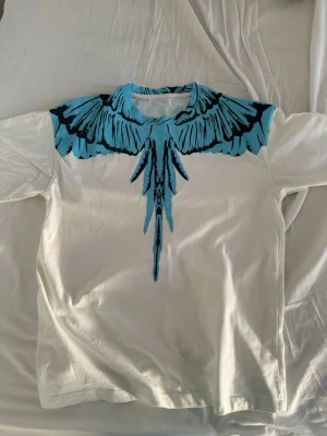 Vit t-shirt med blått grafiskt tryck - Säljer en vit t-shirt med lång ärm och ett stort blått och svart grafiskt tryck som liknar fjädrar runt halsen, både fram och bak. T-shirten har en relaxed passform och är tillverkad i mjuk bomull. Perfekt för dig som vill sticka ut med en unik design. Storlek XL men passar som L