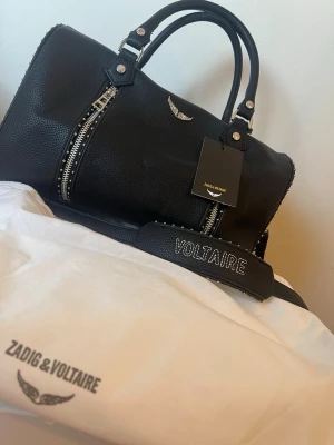 Svart handväska Zadig & Voltaire Medium - Snygg svart handväska från Zadig & Voltaire i strukturerat läder med silverfärgade dragkedjor och detaljer. Väskan har dubbla handtag, avtagbar axelrem med VOLTAIRE-text och ikonisk vinge framtill. Perfekt för dig som gillar en edgy och trendig look.