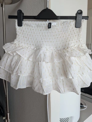 Vit volangkjol från H&M Divided XS - Supersöt vit kortkjol från H&M Divided med flera volanger och små guldiga detaljer. Kjolen har smock i midjan och shorts inuti (bild 3) Stlk XS i jättefint skick.