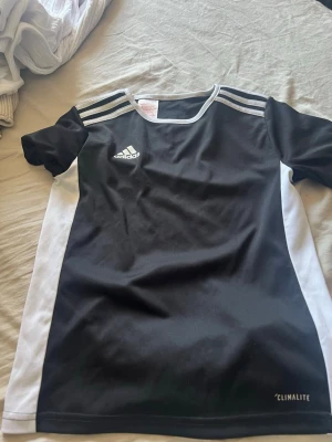 Svart och vit Adidas sporttröja - Säljer en svart och vit sporttröja från Adidas med CLIMALITE-material som andas. Tröjan har korta ärmar, vita ränder på axlarna och vita sidopaneler. Perfekt för träning eller match. Adidas-logga på bröstet.