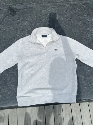 Grå halvzip sweatshirt Lexington - Snygg ljusgrå sweatshirt från Lexington med halv dragkedja och ståkrage. Tröjan har en broderad logga på bröstet och är gjord i mjukt material, perfekt för en avslappnad stil och till skolstarten!