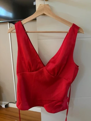 Röd croppad linnetopp - Snygg röd croppad linnetopp med djup v-ringning och breda axelband. Toppen har en figurnära passform och knytband i sidan för extra detalj. Perfekt för dig som vill sticka ut lite extra.