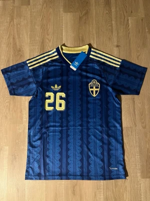 Taha Ali Sverige vm tröja S - Snygg blå fotbollströja från Adidas med gula detaljer, nummer 26 och namnet Ali på ryggen. Tröjan har Sveriges landslagsemblem på bröstet och klassiska Adidas-ränder på axlarna. Tillverkad i lätt och ventilerande polyester.