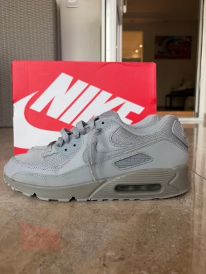 Nike Air Max 90 grå sneakers - Snygga Nike Air Max 90 sneakers i helgrå färg. Skorna har en klassisk siluett med mesh och syntetläder på ovandelen, samt den ikoniska Air Max-dämpningen i sulan. Perfekta för dig som gillar stilrena och sportiga sneakers.
