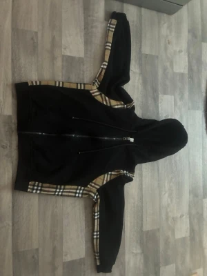 Svart Burberry hoodie  - Säljer en svart hoodie från Burberry i storlek M. Hoodien har dragkedja framtill, klassiska Burberry-rutiga detaljer längs ärmar och sidor samt en stor huva. Tillverkad i mjukt material som känns skönt mot huden. Perfekt för dig som vill ha en stilren men ändå unik look.