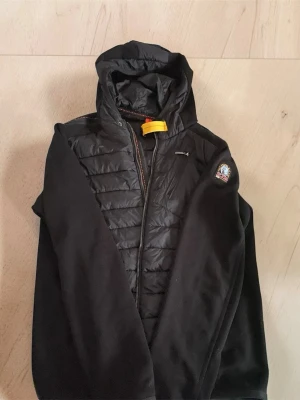 svart parajumper cardigan  - Svart cardiganjacka med huva och quiltad front. Jackan har dragkedja, en bröstficka med dragkedja och en patch på ärmen. Materialet är en mix av vadderad front och mjuka ärmar. Perfekt för lager-på-lager och streetwear.