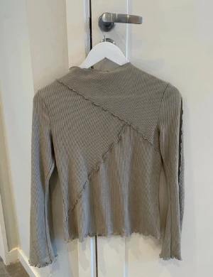 Beige ribbad långärmad topp - Trendig beige långärmad topp i ribbat material med vågiga sömmar och asymmetriska detaljer. Figurnära passform och stretchigt tyg som ger en cool och avslappnad look. Perfekt att styla med jeans eller kjol för en snygg vardagsoutfit.