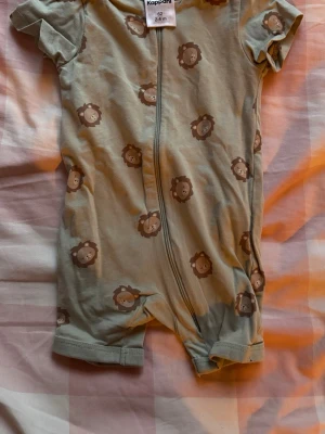 Sött babyset med lejon och väst - Tvådelat babyset med kortärmad body i ljusgrönt med gulliga lejontryck och dragkedja framtill samt ett par beige byxor med resår i midjan och dekorfickor. Perfekt för små barn som vill vara både bekväma och stiliga.