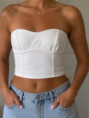Vit axelbandslös korsettopp - Trendig vit korsettopp med bandeau-modell och markerade sömmar. Toppen är axelbandslös och croppad, vilket ger en modern och stilren look. Perfekt att matcha med höga jeans eller kjol för en snygg outfit.