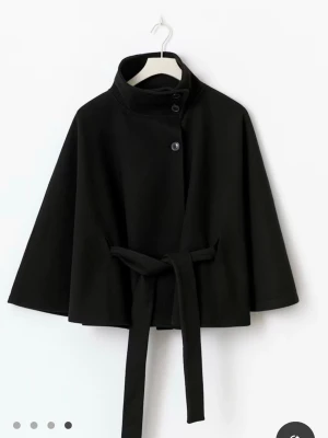 Svart cape-kappa med knytband - Stilren svart cape-kappa med hög krage, knappar framtill och knytband i midjan. Kappan har vida ärmar och en lös passform som ger en cool och modern look. Perfekt för lager-på-lager och passar till många outfits. Kan gå ner i pris vid snabb beställning