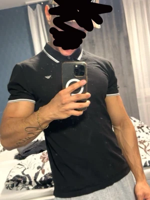 Svart pikétröja från Emporio Armani - Snygg svart pikétröja från Emporio Armani med vit logga på bröstet och vita detaljer på krage och ärmslut. Klassisk passform med korta ärmar och knappar vid halsen. Perfekt för en stilren och avslappnad look.