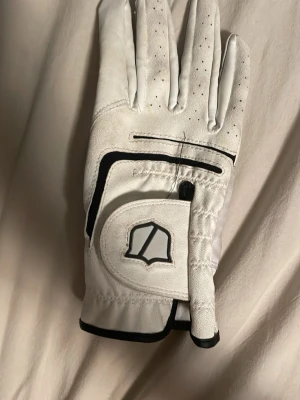 Vit golfhandske från Wilson (höger handske) - (OBS! Kan tvättas innan om man vill) det Säljer en vit golfhandske från Wilson med svarta detaljer och perforeringar för ventilation. Handsken har kardborrestängning och förstärkta partier i handflatan för extra grepp. Perfekt för dig som vill ha stil och funktion på golfbanan.