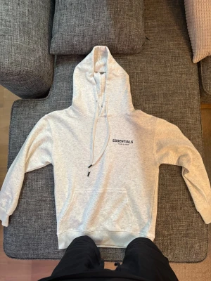 Grå Essentials Fear of God hoodie - Säljer en ljusgrå hoodie från Essentials Fear of God med svart logga på bröstet och stor text på ryggen. Hoodien har huva med dragsko, magficka och ribbade muddar. Perfekt för en clean och avslappnad streetwear-look.
