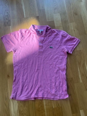 Rosa slim fit pikétröja från Lacoste - Säljer en klassisk rosa pikétröja från Lacoste med korta ärmar och krage. Tröjan har två knappar framtill och den ikoniska krokodilloggan på bröstet. Perfekt för dig som gillar en sportig och stilren look.