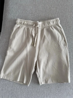 Beige shorts med snörning - Säljer ett par stilrena beige shorts med elastisk midja och snörning framtill. Shortsen har sidofickor och en bakficka, perfekta för chill dagar. Tillverkade i mjukt bomullsliknande material för maximal komfort.