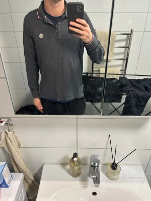 Mörkgrå långärmad piké Moncler - Snygg mörkgrå långärmad pikétröja från Moncler med klassisk krage och knappar framtill. Tröjan har Moncler-logga på bröstet och diskreta röda och vita detaljer på kragen. Perfekt för en stilren och avslappnad look. Priset går att diskuteras vid snabb affär! Storlek M