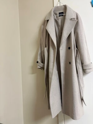 Ljusbeige lång kappa med bälte - Stilren ljusbeige kappa i lång modell med breda slag, knytskärp i midjan och dubbelknäppning framtill. Kappan har knappar vid ärmsluten och en klassisk, rak passform. Perfekt för dig som vill ha en tidlös och trendig look.