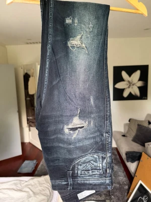 Mörkblå slitna jeans med slitningar - Säljer ett par mörkblå jeans med slitningar och hål på låret och knät. Jeansen har en klassisk femficksmodell och raka ben. Materialet är denim med tvättad look och detaljerade slitningar för en trendig vibe.