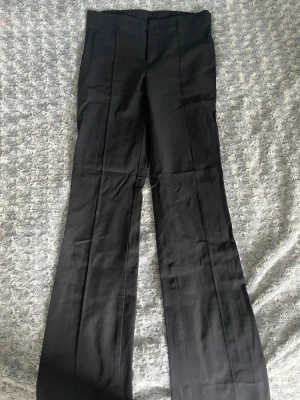Svarta utsvängda kostymbyxor - Snygga svarta kostymbyxor med medelhög midja och utsvängda ben. Byxorna har pressveck framtill för en clean look och stängs med dold knapp och dragkedja. Perfekta för dig som gillar en stilren och trendig vibe.