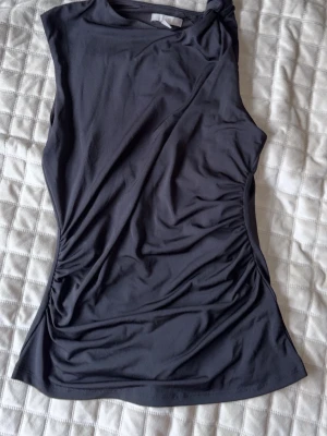 Svart ärmlös topp från H&M - Snygg svart ärmlös topp från H&M med knut vid ena axeln. Toppen har en slät och mjuk yta i stretchigt material, perfekt för en stilren look. Passar till många olika outfits och är enkel att matcha.