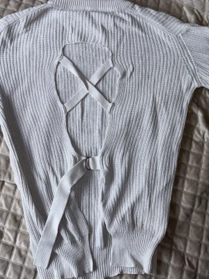 Vit stickad tröja med öppen rygg - Trendig vit stickad tröja med lång ärm och ribbade muddar. Baksidan har en öppen design med dekorativa korsade band. Perfekt för dig som vill ha en unik detalj på din outfit.