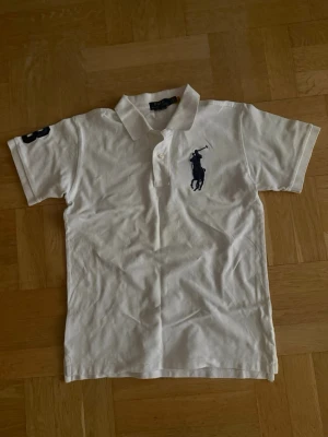 Vit pikétröja med blå logga - Vit pikétröja med klassisk krage och knappar framtill. Tröjan har en mörkblå broderad logga på bröstet och en siffra på ärmen. Kortärmad och tillverkad i mjuk bomull, perfekt för en avslappnad och stilren look.