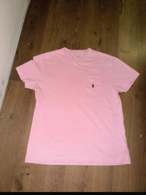 Rosa t-shirt med ficka - Säljer en ljusrosa t-shirt med rund hals och korta ärmar. T-shirten har en bröstficka med en liten broderad logga i mörkblått. Perfekt basic-plagg för dig som gillar enkel och stilren design.