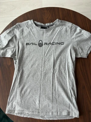 Grå Sail Racing t-shirt - Snygg grå t-shirt från Sail Racing med svart logga tryckt över bröstet. Klassisk rund halsringning med svart och vit rand på insidan av kragen. Perfekt till jeans eller shorts för en avslappnad stil.