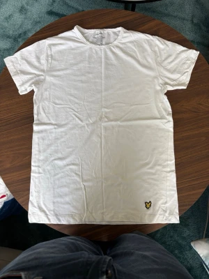 Vit t-shirt från Lyle & Scott - Klassisk vit t-shirt från Lyle & Scott med rund hals och korta ärmar. Tillverkad i mjuk bomull med lite elastan för extra stretch. Liten gul logga med örn broderad nere vid fållen. Perfekt basic-plagg till garderoben.