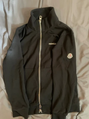 Svart zip-hoodie från Moncler - Svart hoodie med dragkedja från Moncler. Klassisk design med hög krage, broderad Moncler-logga på ärmen och vit Moncler-text på bröstet. Mjuk bomullskänsla och ribbade muddar. Perfekt för en stilren och avslappnad look.