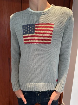 Polo Ralph Lauren Flag Sweater – Storlek S - Polo Ralph Lauren-tröja i superfint skick    Märke: Polo Ralph Lauren  Modell: Klassisk stickad tröja med USA-flagga  Storlek: XL för ungdom/junior (motsvarar en vuxen Small)  Passform: Modellen på bilderna är 182 cm lång för att visa hur den sitter  Material: 100% bomull  Skick: Superfint skick, inga anmärkningar  Äkthet: 100% äkta – tröjan är köpt via auktoriserad återförsäljare och har korrekta inner- och nacketiketter (samt den officiella märkningen för material och tillverkning)  